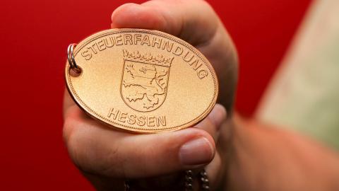 Eine Hand hält die Plakette der hessischen Steuerfahndung ins Bild.