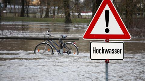 Bild eines Hochwassergebiets mit einem Schild, das vor Hochwasser warnt. Im Bildhintergrund steht ein Fahrad halb versunken im Wasser.