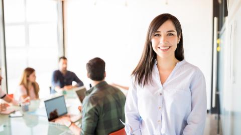 Im Bildvordergrund steht eine junge Frau, im Bildhintergrund sitzt ein Team am Konferenztisch.