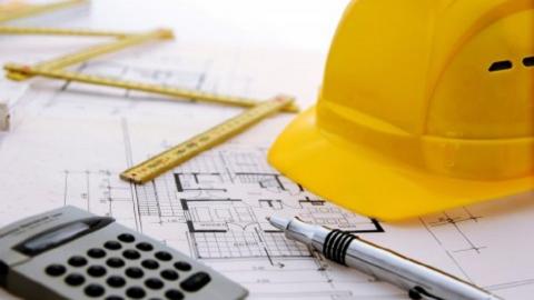 Zu sehen sind ein Taschenrechner, ein Kugelschreiber, ein Schutzhelm sowie ein Lineal auf einem gezeichneten Bauplan.