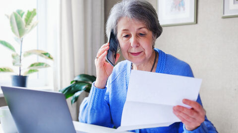 Eine Frau sitzt mit einem Schriftstück in der Hand vor ihrem Notebook und telefoniert.