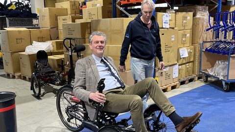 Finanzstaatssekretär Dr. Worms sitzt in einem Velobike und testet es in der Fertigungshalle, neben ihm Alexander Kraft, Pressesprecher des Unternehmens.