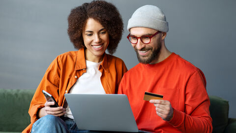 E-Payment macht es möglich, einfach und schnell mit Smartphone oder Laptop Bezahlungen abzuwickeln. Eine Frau und ein Mann sitzen auf einer Couch und bezahlen online mit Smartphone und Laptop.