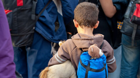 Hessen hilft Geflüchteten. Im Bildvordergrund zu sehen ist ein kleiner Junge mit Kuscheltier und Rucksack, im Bildhintergrund Erwachsene.
