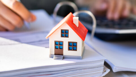 Das Modell eines Hauses steht auf einem Aktenordner. Im Hintergrund tippt jemand Zahlen in einen Taschenrechner ein und füllt ein Formular aus.
