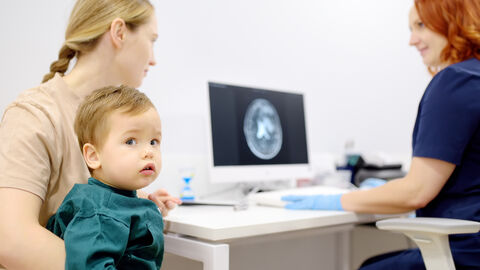 Untersuchung bei einer Kinderneurologin. Ein Kind ist mit seiner Mutter bei einer neurologischen Untersuchung bei einer Ärztin, die auf einen Bildschirm mit einem MRT-Scan blickt.