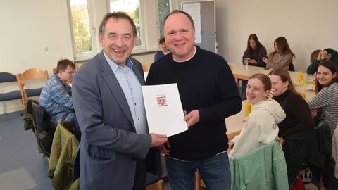 Hessens Finanzminister Professor Dr. R. Alexander Lorz überreicht den Förderbescheid an den Pfarrer der Gemeinde St. Birgid, Frank Schindling. Im Bildhintergrund sind Firmandinnen und Firmanden zu sehen.