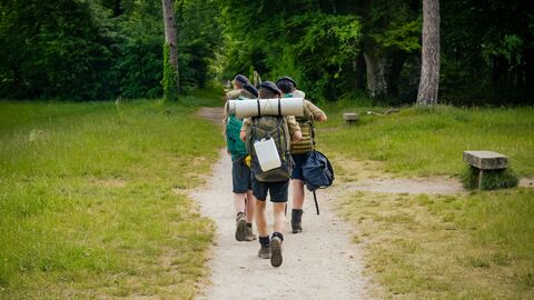 Vier Männer als Pfadfinder gekleidet und mit Rucksack auf dem Rücken sind von hinten zu sehen und gehen einen Pfad entlang.