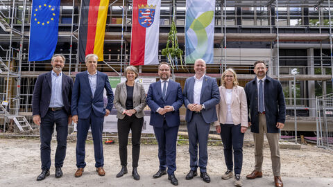 Mehrere Menschen stehen vor Baustelle mit Gerüst. Am Gerüst hängen vier Flaggen.