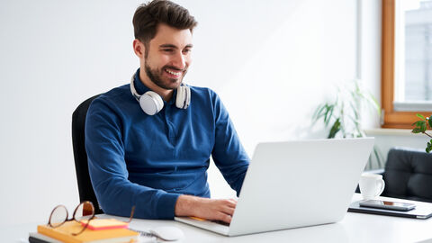 Ein Mann tippt an einem Laptop. Neben ihm liegen Bücher und eine Brille.