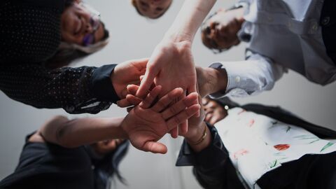 Fünf Menschen legen als Zeichen ihres Zusammenhalts ihre Hände aufeinander. Die Hände sind von unten zu sehen.