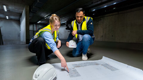 Eine Frau und ein Mann in Schutzkleidung beugen sich gemeinsam auf einer Baustelle über einen Plan mit einem Grundriss.